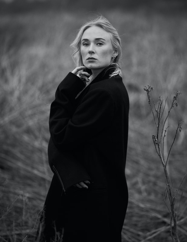 Blonde Swedish girl and model Maija standing in the wind by the beach dressed in dark coat. Black and white photography by Swedish photographer Tomas Lissåker. 