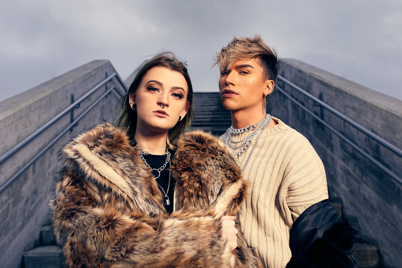 One female and one male model posing in front of concrete stairs dressed in beige, brown and black street fashion and looking straight into the camera. 
